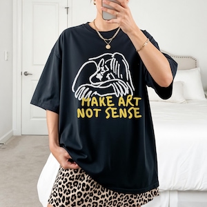 Puede incluir: Camiseta negra de gran tamaño con un gráfico blanco y amarillo que dice "MAKE ART NOT SENSE". El gráfico presenta un dibujo lineal de una persona abrazándose. La persona lleva una falda con estampado de leopardo.