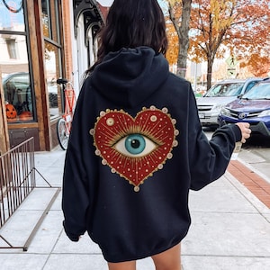 May include: Black hoodie featuring a large heart-shaped design on the back. The heart is red with a detailed eye in the center, surrounded by gold accents and white dots. The hoodie is a casual garment.