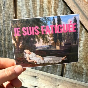 May include: A sticker with a painting of a woman lying down in a field. The text "JE SUIS FATIGU&Eacute;E" is printed in pink on the sticker.