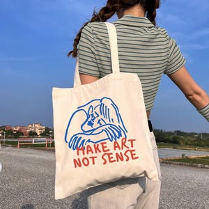 Może przedstawiać: Kremowa torba typu tote z niebiesko-czerwoną grafiką. Grafika przedstawia rysunek osoby i napis "MAKE ART NOT SENSE" w kolorze czerwonym. Torba ma kremowy pasek.