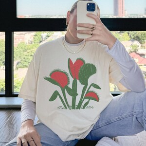 May include: Cream-colored t-shirt featuring a graphic of a Venus flytrap plant with red and green details. The shirt is paired with a long-sleeved white shirt and blue jeans. The person is holding a phone.