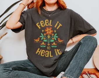 Puede incluir: Una camiseta gris oscuro con un diseño gráfico que presenta una flor con pájaros y el texto "Feel it to heal it".