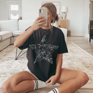 May include: Black t-shirt featuring a distressed white star graphic. The short-sleeved shirt is worn with white shorts and white socks with black stripes. The person is sitting on a rug.