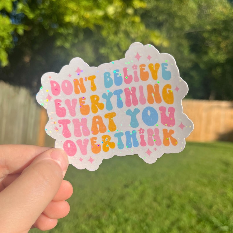 Op de afbeelding: Een witte sticker met een regenboogkleurige glitterachtergrond en de tekst "Geloof niet alles wat je overdenkt" in een kleurrijk, retro lettertype.