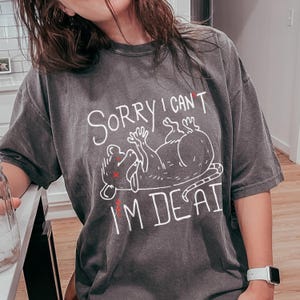 May include: A dark grey t-shirt with a white graphic of a rat lying on its back with the text "Sorry I can't I'm dead".