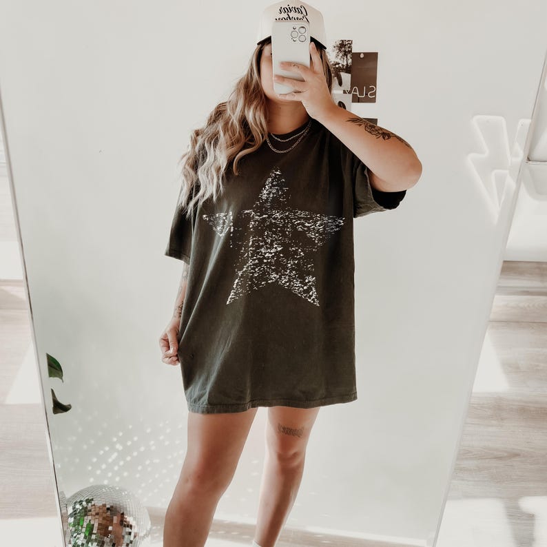 May include: An olive green oversized t-shirt with a large, distressed white star graphic. The person is wearing a white baseball cap with black text. The shirt is paired with a silver necklace.