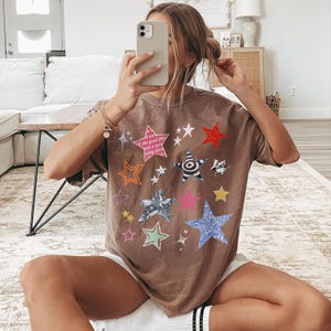 Comfort Colors 1717 T- Shirt
Color: Espresso (brown)
Graphic: bold star graphic design in multiple colors and textures. Features a mix of embroidered, metallic, and printed stars, along with abstract celestial details.