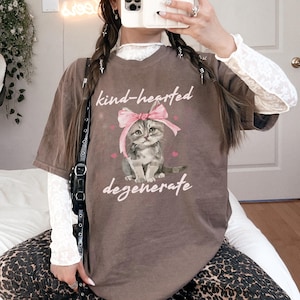Comfort Colors 1717 Shirt
Color: Espresso Brown
Graphic: Illustration of a gray tabby kitten sitting with a large pink bow tied on its head. The kitten is surrounded by small pink hearts and framed by the handwritten-style phrase "kind-hearted degenerate" in soft, playful lettering.
keywords: cutecore, funny cat mom gift, cat lady, cute kitten, pet parent