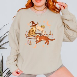 May include: Beige sweatshirt featuring a whimsical illustration of three cats. One cat wears a witch hat, another a floral crown, and the third a crown. The design includes candles and crescent moons, creating a Halloween or mystical theme.