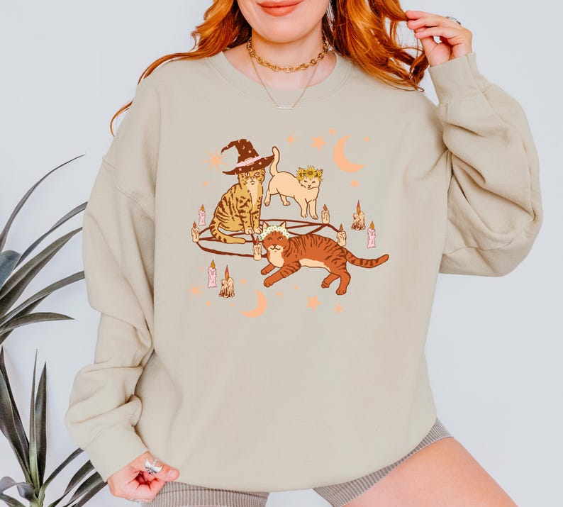 May include: Beige sweatshirt featuring a whimsical illustration of three cats. One cat wears a witch hat, another a floral crown, and the third a crown. The design includes candles and crescent moons, creating a Halloween or mystical theme.