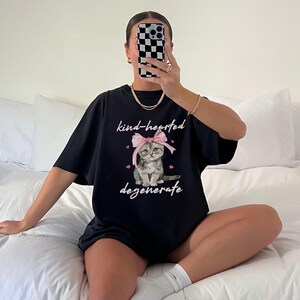 Comfort Colors 1717 Shirt
Color: Black
Graphic: Illustration of a gray tabby kitten sitting with a large pink bow tied on its head. The kitten is surrounded by small pink hearts and framed by the handwritten-style phrase "kind-hearted degenerate" in soft, playful lettering.