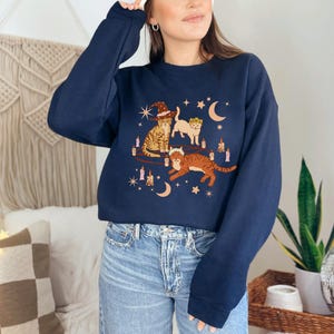 May include: A navy blue sweatshirt featuring a whimsical illustration of three cats. One cat wears a witch hat, another a crown, and the third is leaping. The design includes candles, moons, and stars.