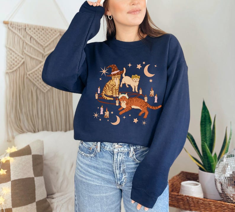May include: A navy blue sweatshirt featuring a whimsical illustration of three cats. One cat wears a witch hat, another a crown, and the third is leaping. The design includes candles, moons, and stars.