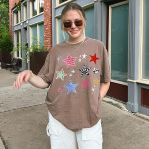 Comfort Colors 1717 T-Shirt
Color: Espresso Brown
Graphic: Colorful collage-style star graphic featuring various hand-drawn and patterned stars scattered across the chest. The stars include a mix of animal prints, stripes, solid colors, and textures, with some metallic and doodle-like accents.