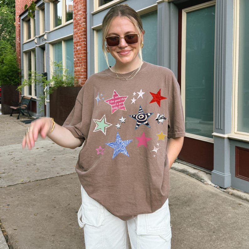 Comfort Colors 1717 T-Shirt
Color: Espresso Brown
Graphic: Colorful collage-style star graphic featuring various hand-drawn and patterned stars scattered across the chest. The stars include a mix of animal prints, stripes, solid colors, and textures, with some metallic and doodle-like accents.