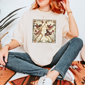 Pu&ograve; includere: Una t-shirt color crema con un'illustrazione in stile vintage di due fate che danzano in un giardino. Le fate indossano abiti rossi e verdi e hanno capelli e ali marroni. Lo sfondo &egrave; un motivo floreale con una combinazione di colori marrone e verde.