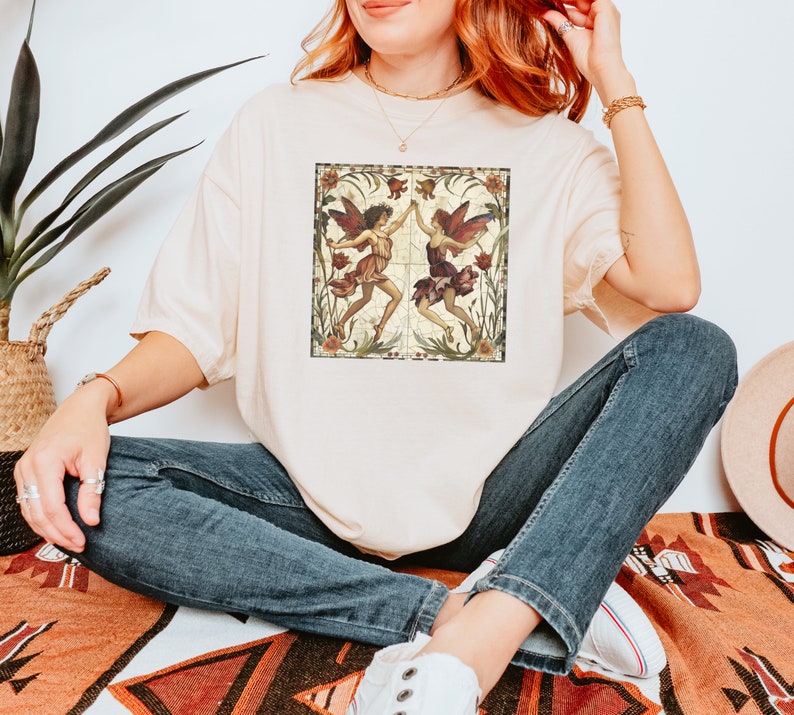 Pu&ograve; includere: Una t-shirt color crema con un'illustrazione in stile vintage di due fate che danzano in un giardino. Le fate indossano abiti rossi e verdi e hanno capelli e ali marroni. Lo sfondo &egrave; un motivo floreale con una combinazione di colori marrone e verde.