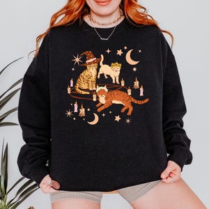 May include: Dark charcoal sweatshirt featuring a whimsical illustration of three cats. One cat wears a witch hat, another a crown, and the third a floral crown. The design includes candles, moons, and stars, creating a mystical theme.
