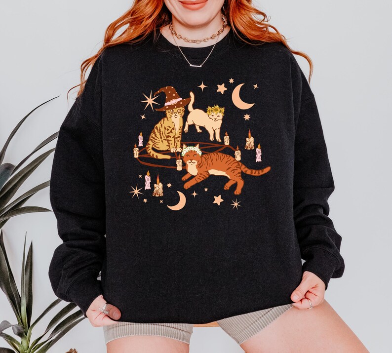 May include: Dark charcoal sweatshirt featuring a whimsical illustration of three cats. One cat wears a witch hat, another a crown, and the third a floral crown. The design includes candles, moons, and stars, creating a mystical theme.