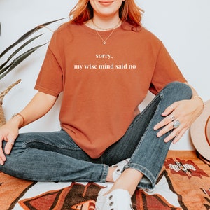 May include: A woman wearing a rust-colored t-shirt with the text "sorry, my wise mind said no" in white. She is sitting on a patterned rug with her legs crossed and wearing blue jeans and white sneakers.