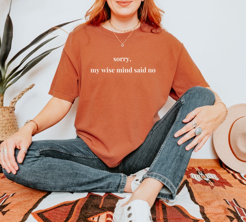 May include: A woman wearing a rust-colored t-shirt with the text "sorry, my wise mind said no" in white. She is sitting on a patterned rug with her legs crossed and wearing blue jeans and white sneakers.