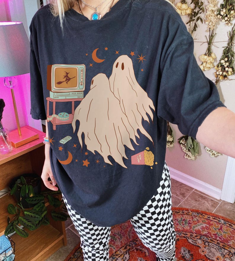 Comfort Colors 1717 Shirt
Color: Black
Graphic: Two cartoon-style ghosts watch a witch flying on a TV screen surrounded by stars, popcorn, VHS tapes, and crescent moons. The illustration has a warm vintage Halloween palette on a cozy crewneck sweatshirt.