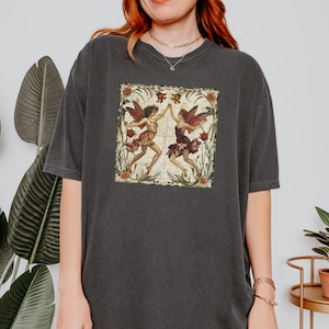 Pu&ograve; includere: Una maglietta oversize di colore grigio scuro con un'illustrazione vintage di due fate con le ali, stampata sul davanti. Le fate sono circondate da fiori e foglie.