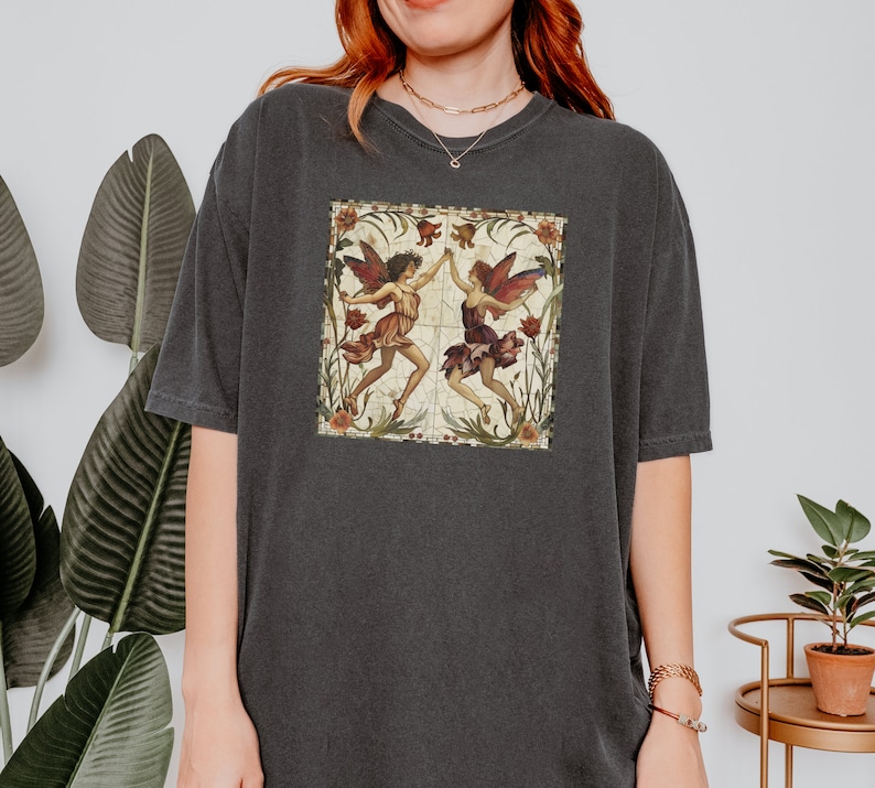 Pu&ograve; includere: Una maglietta oversize di colore grigio scuro con un'illustrazione vintage di due fate con le ali, stampata sul davanti. Le fate sono circondate da fiori e foglie.