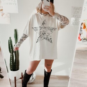 May include: Cream-colored oversized t-shirt with a large, distressed black star graphic. The shirt is paired with sheer patterned sleeves, black boots, and a black baseball cap. A cactus and mirror are in the background.