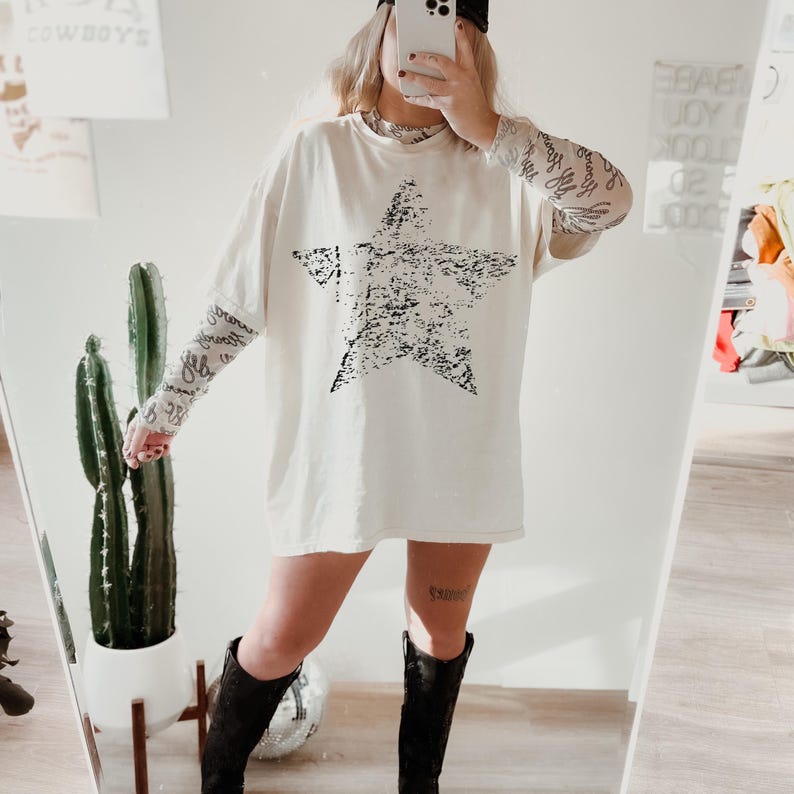 May include: Cream-colored oversized t-shirt with a large, distressed black star graphic. The shirt is paired with sheer patterned sleeves, black boots, and a black baseball cap. A cactus and mirror are in the background.