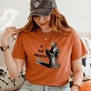 Op de afbeelding: Een vrouw draagt een oranje T-shirt met een afbeelding van de Grim Reaper die "It has pockets!" zegt. De Grim Reaper houdt de rand van zijn gewaad omhoog om de zakken te laten zien. De vrouw draagt ook een zwarte baseballpet met de woorden "Stay Spooky" erop geborduurd.