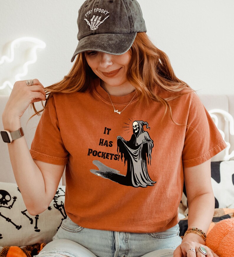 Op de afbeelding: Een vrouw draagt een oranje T-shirt met een afbeelding van de Grim Reaper die "It has pockets!" zegt. De Grim Reaper houdt de rand van zijn gewaad omhoog om de zakken te laten zien. De vrouw draagt ook een zwarte baseballpet met de woorden "Stay Spooky" erop geborduurd.