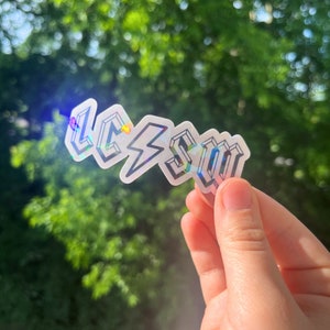 May include: A holographic sticker with the word "L.C.S.U" in a bold, stylized font. The sticker is iridescent and reflects light in a rainbow of colors.