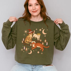 May include: Olive green sweatshirt featuring a whimsical illustration of three cats adorned with witch hats and floral crowns. The cats are surrounded by candles, stars, and crescent moons. The sweatshirt is a comfortable, casual style.