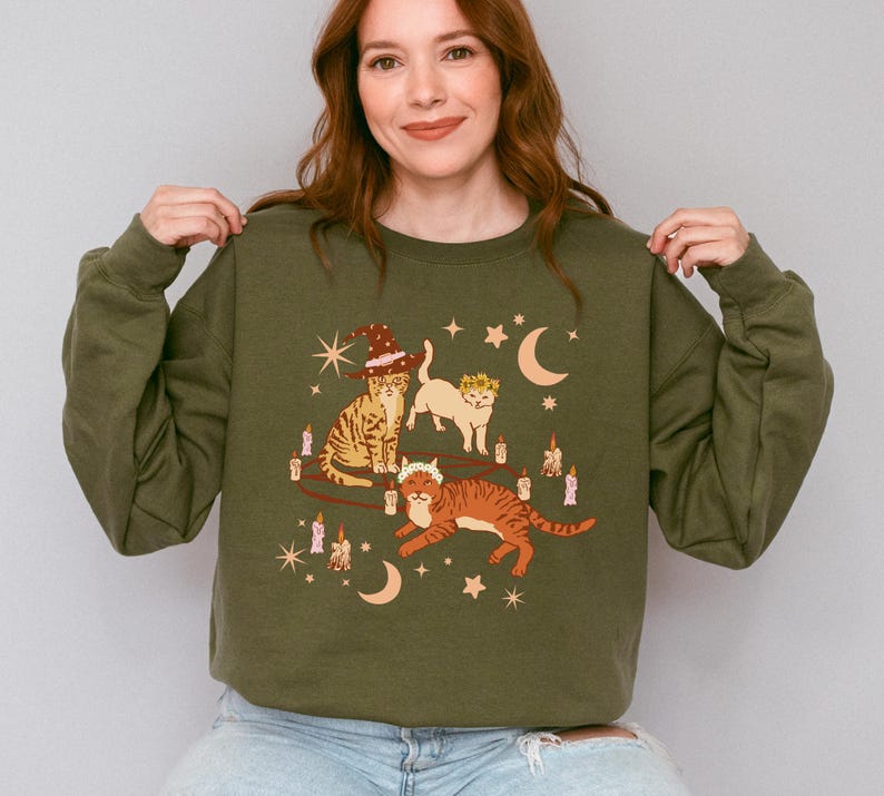 May include: Olive green sweatshirt featuring a whimsical illustration of three cats adorned with witch hats and floral crowns. The cats are surrounded by candles, stars, and crescent moons. The sweatshirt is a comfortable, casual style.