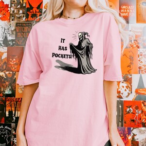 Op de afbeelding: Roze T-shirt met een afbeelding van een skelet in een gewaad met de tekst "It has pockets!"