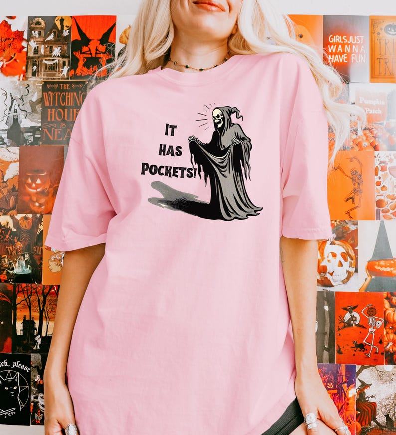 Op de afbeelding: Roze T-shirt met een afbeelding van een skelet in een gewaad met de tekst "It has pockets!"
