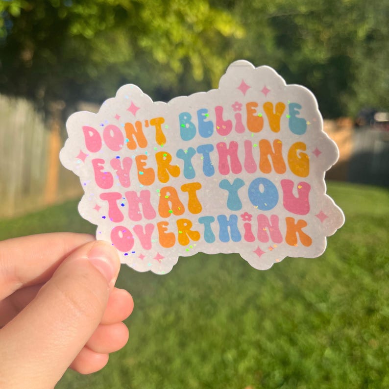 Op de afbeelding: Een witte sticker met regenboogkleurige tekst die "Geloof niet alles wat je overdenkt" zegt.