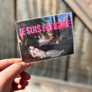 May include: A sticker with a painting of a woman lying down in a field. The text "JE SUIS FATIGU&Eacute;E" is printed in pink on the sticker.