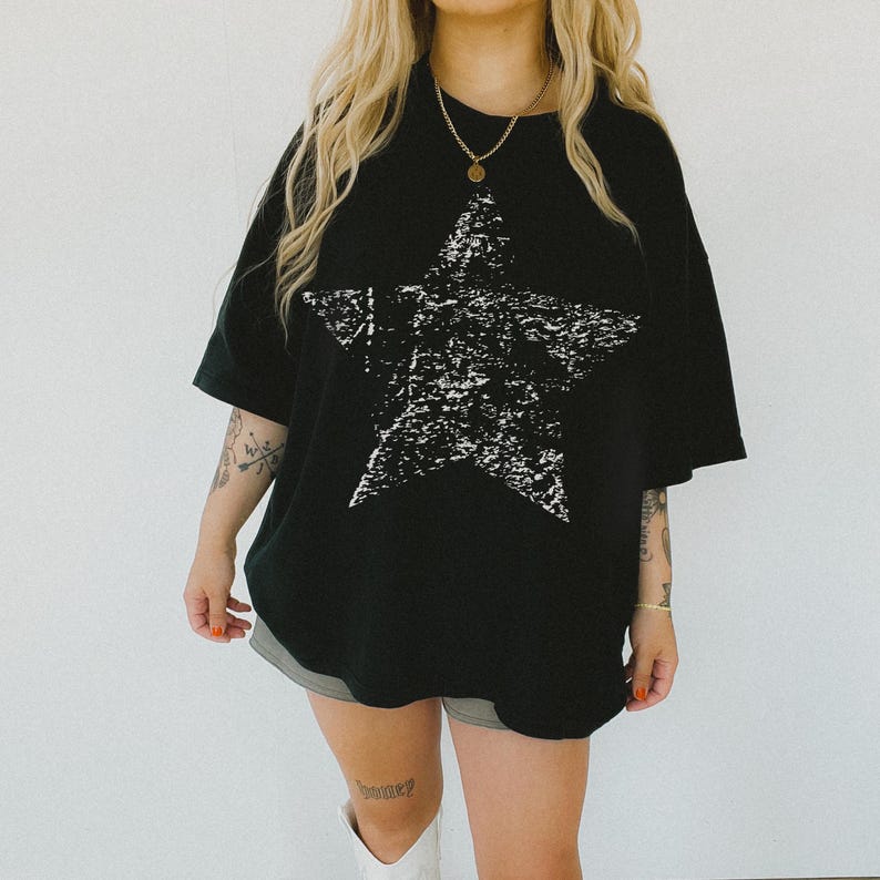 May include: Black oversized t-shirt featuring a large, distressed white star graphic. The shirt has short sleeves and a relaxed fit. The model is wearing the shirt with shorts and boots.