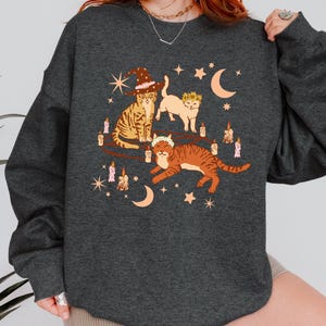 May include: Dark gray sweatshirt featuring a whimsical illustration of three cats. One cat wears a witch hat, another a floral crown, and the third a floral crown. The design includes candles, stars, and crescent moons, creating a magical theme.
