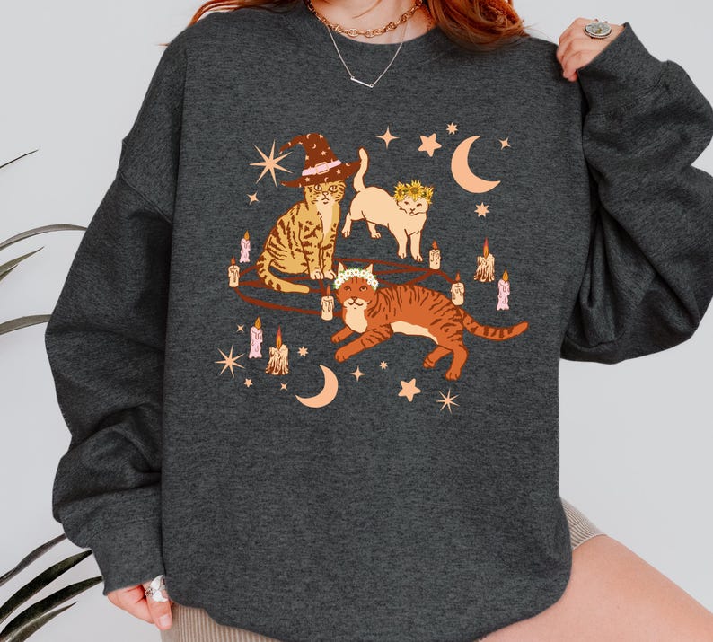 May include: Dark gray sweatshirt featuring a whimsical illustration of three cats. One cat wears a witch hat, another a floral crown, and the third a floral crown. The design includes candles, stars, and crescent moons, creating a magical theme.