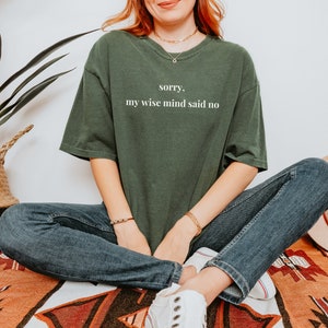May include: A woman wearing a dark green t-shirt with the text "sorry, my wise mind said no" in white lettering. She is wearing blue jeans and white sneakers.