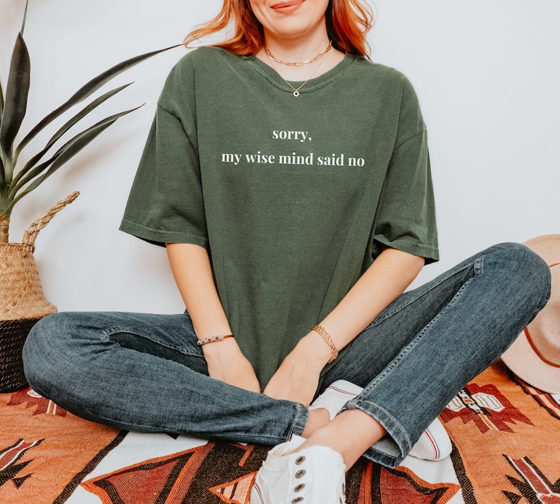 May include: A woman wearing a dark green t-shirt with the text "sorry, my wise mind said no" in white lettering. She is wearing blue jeans and white sneakers.