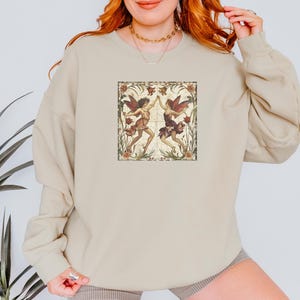 May include: A beige sweatshirt with a vintage-style illustration of two fairies dancing in a floral frame. The fairies are wearing flowing dresses and have wings. The frame is surrounded by a border of flowers and leaves.