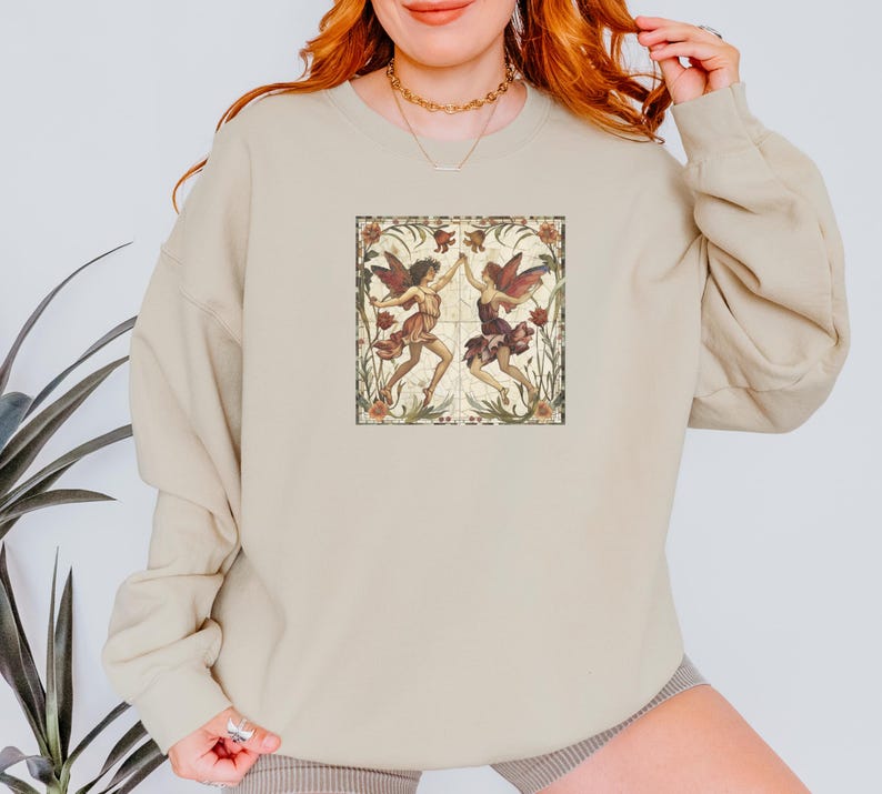 May include: A beige sweatshirt with a vintage-style illustration of two fairies dancing in a floral frame. The fairies are wearing flowing dresses and have wings. The frame is surrounded by a border of flowers and leaves.