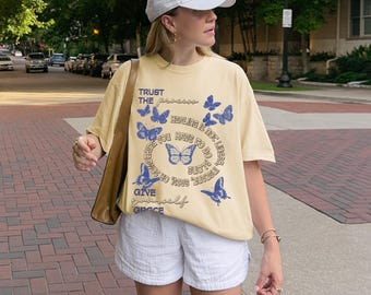 May include: A pale yellow t-shirt featuring blue butterfly designs and the text "TRUST THE process, HEALING IS not linear, YOU HAVE TO DO alone, GIVE yourself GRACE". The shirt is worn with white shorts and a white baseball cap.