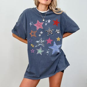 Comfort Colors 1717 T- Shirt
Color: Denim
Graphic: bold star graphic design in multiple colors and textures. Features a mix of embroidered, metallic, and printed stars, along with abstract celestial details.
