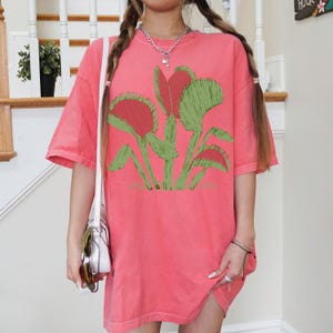 May include: A pink oversized t-shirt with a graphic print of a Venus flytrap plant. The shirt has a relaxed fit and short sleeves.