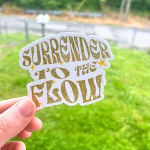K&ouml;nnte beinhalten: Ein wei&szlig;er Aufkleber mit goldener Schrift, auf dem "Surrender to the Flow" steht, mit kleinen goldenen Sternen um den Text herum.